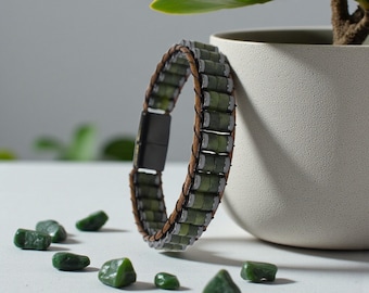 Può includere: Un braccialetto di giada verde con chiusura magnetica. Il braccialetto presenta perline di giada rettangolari, una fascia in pelle marrone e una chiusura magnetica nera. Diverse pietre di giada grezza sono sparse intorno al braccialetto.