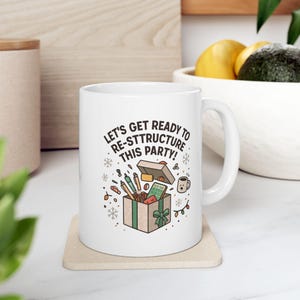 May include: White ceramic mug with a playful design. The mug displays the text "LET'S GET READY TO RE-STTRUCTURE THIS PARTY!" and illustrations of stationery and a gift box. The mug sits on a beige coaster.