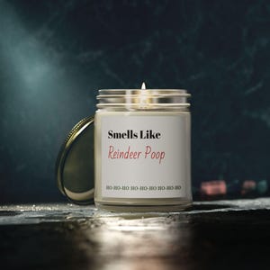 May include: A lit candle in a clear glass jar with a silver lid. The label reads "Smells Like Reindeer Poop" in black and red text, with "HO-HO-HO" repeated below. The jar is on a dark surface.