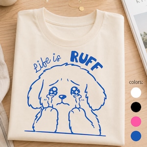 Könnte beinhalten: Cremefarbenes T-Shirt mit einem blauen Cartoon-Hund mit Tränen in den Augen und dem Schriftzug "Life is Ruff". Das Design ist mittig platziert. Das T-Shirt liegt gefaltet auf einer Holzoberfläche. Farben: Weiß, Schwarz, Pink und Blau.