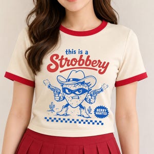 May include: A cream-colored ringer t-shirt with red trim at the neck and sleeves. The graphic features a strawberry character wearing a cowboy hat and mask, holding two pistols, with the text "this is a Strobbery" and "BERRY WANTED".
