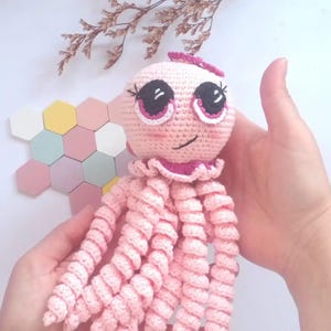 Pode incluir: Boneca de polvo rosa de crochê feita à mão com olhos grandes e expressivos e tentáculos encaracolados. A boneca tem um pequeno laço rosa na cabeça e uma gola com babados. O fundo inclui um arranjo decorativo de formas geométricas em tons pastel e flores secas.