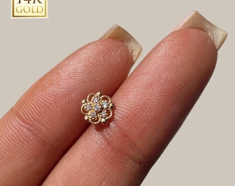 Könnte beinhalten: Ein kleiner, goldfarbener Blumen-Ohrstecker mit klaren Edelsteinen. Der Ohrring wird zwischen zwei Fingern gehalten. In der oberen linken Ecke des Bildes befindet sich ein 14K Goldstempel.