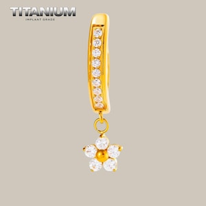 May include: A gold-colored titanium implant grade hoop earring featuring a curved bar set with a vertical row of nine clear round stones and a dangling flower-shaped charm composed of five clear petals surrounding a central gold-colored bead.