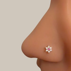 May include: A gold-colored nose stud in the shape of a flower. The flower has six white, round gemstones surrounding a central pink gemstone. The stud is shown on a person's nose.