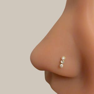 May include: A gold nose stud featuring three clear, round, sparkling gemstones. The gemstones are set in a vertical line, creating a delicate and elegant look. The jewelry is shown on a person's nose.