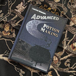 Puede incluir: Un libro de tapa dura titulado "Advanced Potion Making" de Libatius Borage. El libro tiene una cubierta azul oscuro con letras plateadas y un gráfico de un caldero. El fondo es una tela negra con ilustraciones doradas y beige.