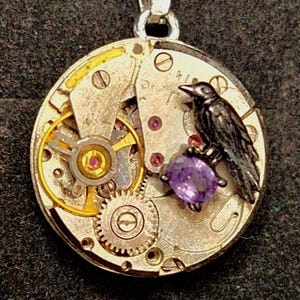 May include: Silver-toned pendant necklace featuring a circular watch mechanism design. A small, black raven figure is perched above a purple gemstone. The pendant hangs from a silver chain, creating a unique, steampunk-inspired accessory.