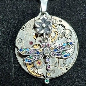 May include: Silver-toned pendant necklace featuring a dragonfly design. The dragonfly has colorful gemstone accents on its wings and body. The pendant hangs from a silver chain. The background of the pendant is a watch mechanism.