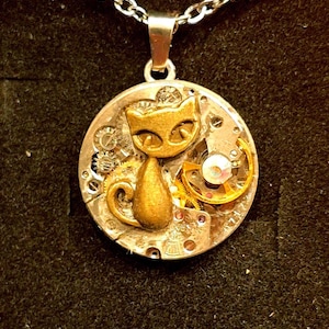May include: A steampunk pendant necklace with a gold cat charm. The round pendant incorporates watch gears and a small crystal. The necklace has a silver chain and clasp, set against a dark background.