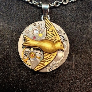 May include: A silver necklace with a pendant. The pendant features a gold-coloured bird design set against a circular background of watch gears. The chain is a delicate silver link.