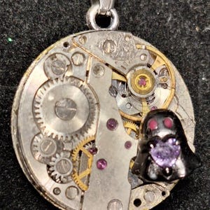 May include: A silver pendant necklace featuring a repurposed watch mechanism. The round pendant showcases intricate gears, ruby accents, and a small, black figure with a purple heart-shaped gem.