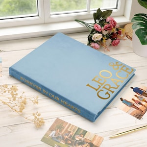May include: A light blue photo album with gold lettering that reads "Leo & Grace" and "Forever in Our Hearts." The album is rectangular and sits on a white surface with photos and flowers nearby.