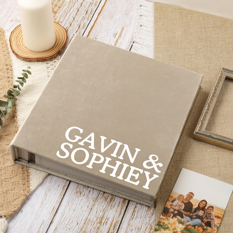 May include: A beige photo album with the names "GAVIN & SOPHIEY" in white lettering. The album is on a wooden surface, with a candle, a framed photo, and a family photo visible.