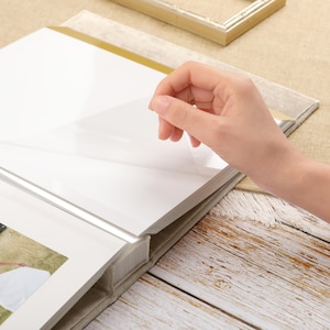 May include: A photo album with white pages and a clear protective sheet being lifted by a hand. The album has a light-colored fabric cover and a gold-colored accent. The background includes a wooden surface and a gold-colored frame.
