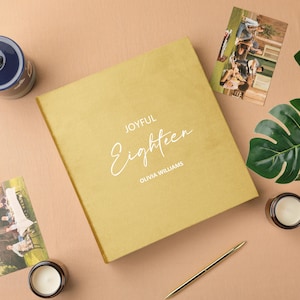 May include: A gold-colored photo album with the words "JOYFUL Eighteen OLIVIA WILLIAMS" in white script. The album is surrounded by photos, candles, a pen, and a decorative leaf on a neutral surface.