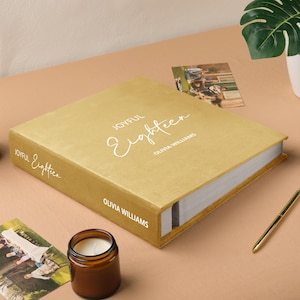 May include: A gold photo album with the words "Joyful Eighteen" and "Olivia Williams" in white script. The album is on a tan surface with a candle, pen, and photos.