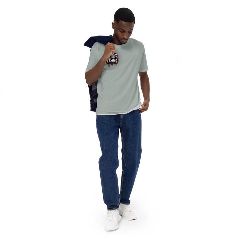 May include: A light green t-shirt with the word "YANKS" and a graphic design on the front. The shirt is paired with blue jeans and white sneakers. A dark blue jacket is draped over the shoulder.