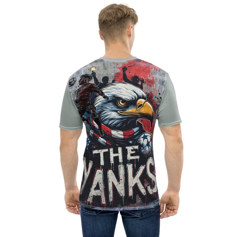 The back features a massive, high-impact illustration. A fierce bald eagle, wrapped in an American flag scarf, dominates the center. To the left, a soccer player leaps into action, capturing the energy of a live match. The background mimics a stadium wall covered in graffiti, complete with silhouettes of fans holding flares, creating an electric atmosphere. The text "THE YANKS" is displayed in a bold, dripping font at the bottom.
The front has smaller, shield-style logo on the left chest.