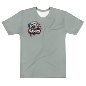 May include: A light gray t-shirt featuring a graphic of an eagle's head with the text "THE YANKS" and a red, white, and blue design. The shirt has short sleeves and a crew neck.