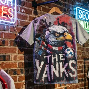 The back features a massive, high-impact illustration. A fierce bald eagle, wrapped in an American flag scarf, dominates the center. To the left, a soccer player leaps into action, capturing the energy of a live match. The background mimics a stadium wall covered in graffiti, complete with silhouettes of fans holding flares, creating an electric atmosphere. The text "THE YANKS" is displayed in a bold, dripping font at the bottom.
The front has smaller, shield-style logo on the left chest.