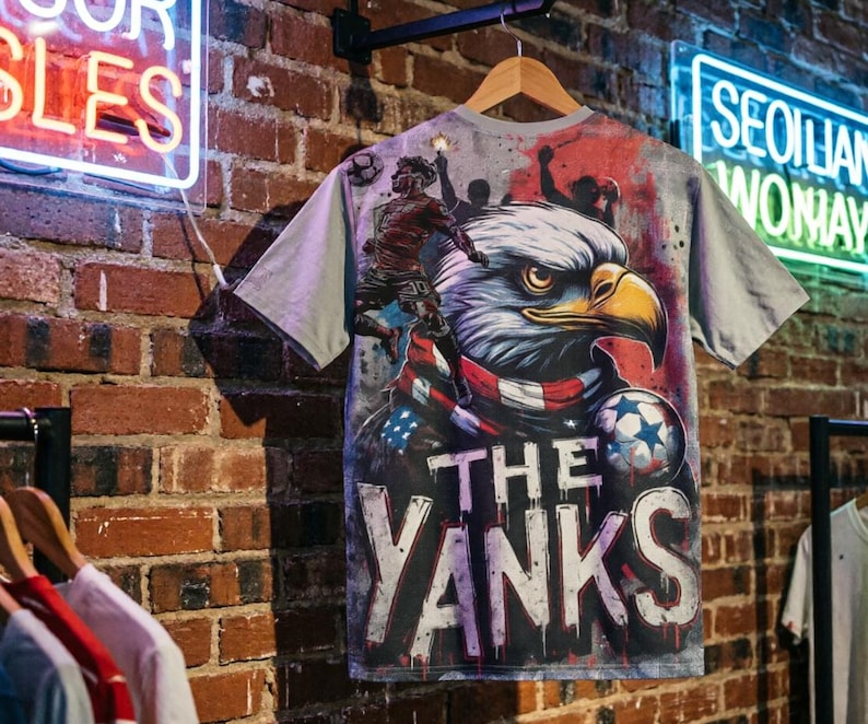 The back features a massive, high-impact illustration. A fierce bald eagle, wrapped in an American flag scarf, dominates the center. To the left, a soccer player leaps into action, capturing the energy of a live match. The background mimics a stadium wall covered in graffiti, complete with silhouettes of fans holding flares, creating an electric atmosphere. The text "THE YANKS" is displayed in a bold, dripping font at the bottom.
The front has smaller, shield-style logo on the left chest.
