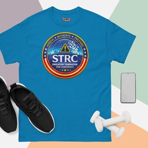 Puede incluir: Una camiseta turquesa con un diseño gráfico circular con el texto "STRC Simulation Termination Risk Conference". El diseño incluye un globo, estrellas y las palabras "Assess, Mitigate, Prepare".