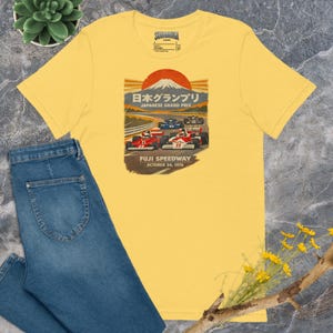 May include: Yellow t-shirt featuring a vintage Japanese Grand Prix design. The graphic includes race cars, a rising sun, and text in Japanese and English, with "Fuji Speedway October 24, 1976". The shirt is paired with blue jeans.