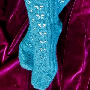Könnte beinhalten: Ein Paar tealblaue Stricksocken mit einem Spitzenmuster. Die Socken liegen auf einem tiefvioletten Samt-Hintergrund.
