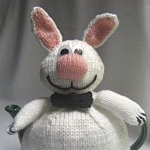 May include: White knitted teapot cosy shaped like a rabbit with pink ears, nose, and a black bow tie.