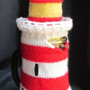 May include: A red and white striped knitted coffee pot cosy with a yellow and red knitted hat on top. The cosy is shaped like a lighthouse.