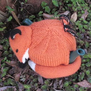 May include: A knitted orange fox teapot cosy with a butterfly design. The cosy is made of soft yarn and has a cute fox face with black eyes and a white nose.