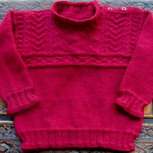 May include: A red knitted sweater with a high neck and a cable knit pattern on the shoulders and sleeves.