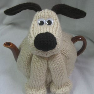 May include: A knitted beige dog tea cosy with brown ears and a black nose. The cosy is on a brown teapot.