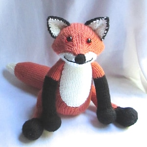 May include: A knitted fox toy with orange fur, white chest, and black legs and feet. The fox has red eyes and a black nose.