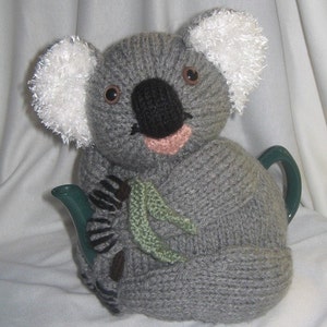 Op de afbeelding: Een grijze gebreide koala thee-cozy met witte vacht oren en bruine ogen. De cozy houdt een groen eucalyptus blad in zijn poten en bedekt een groene theepot.