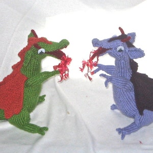 Puede incluir: Dos dragones de punto enfrentados, uno es verde y rojo, el otro es morado y negro. Ambos dragones tienen hilo rojo saliendo de sus bocas.