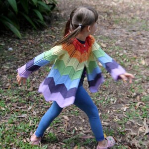May include: A young person wearing a rainbow crocheted cardigan with a chevron pattern. The cardigan is open in the front and has long sleeves. The person is wearing blue jeans and has their hair pulled back in pigtails.