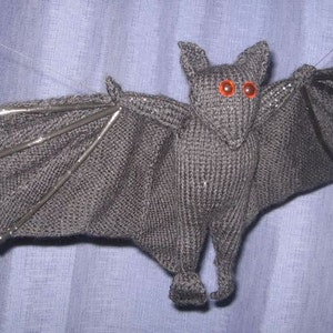 May include: A gray knitted bat with red eyes. The bat has wings with black stitching and is hanging from a string.