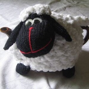 May include: A white knitted teapot cozy shaped like a sheep with a black face, red nose, and large black eyes.