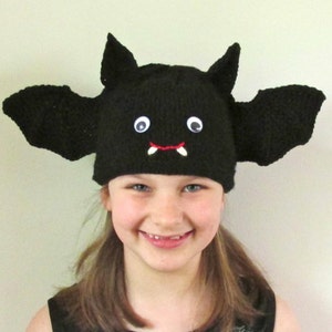 May include: A black knitted bat hat with googly eyes and red yarn fangs. The hat has large bat wings that stick out from the sides. A child is wearing the hat and smiling.