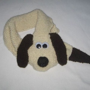 Peut inclure: Une écharpe en tricot en forme de chien avec des oreilles brunes et un nez noir. L'écharpe est beige avec des yeux blancs.