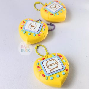 May include: Three yellow heart-shaped keychains with pixelated designs. Each keychain has a small screen with a different graphic, surrounded by colorful sprinkles and a light blue border. The keychains have a gold or purple chain.