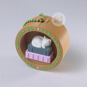 May include: A brown, circular ornament featuring a white cat sleeping on a stack of books. The ornament has a green beaded top and a green leaf design around the edge. The books are gray and purple.