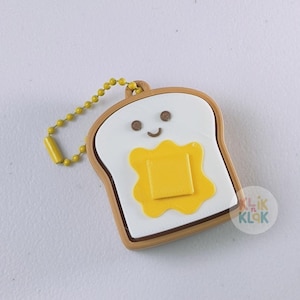 May include: A novelty keychain shaped like a slice of toast with a smiling face and a pat of butter. The keychain has a yellow chain and a yellow bead. The toast is white with brown edges.