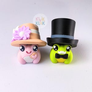 May include: Two charming frog figurines. One is pink with a hat, flower, and heart. The other is green with a top hat and bow tie. The figurines have large eyes and are set against a white background.