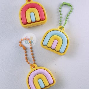 May include: Three rainbow-shaped keyrings with yellow, pink, blue, and yellow arches. Each keyring has a different coloured chain. The keyrings are made of a hard plastic material. The text "Klik Klok" is visible on one of the keyrings.