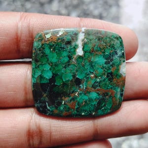 Può includere: Una gemma di forma quadrata con un sorprendente motivo screziato verde e marrone. La superficie lucida mostra un mix di tonalità verde scuro, verde chiaro e marrone. La gemma è tenuta in una mano.