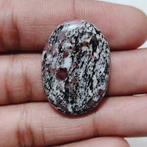 以下が含まれることがあります： 黒、白、ピンクがかった赤色の独特な模様が特徴的な、楕円形の宝石。研磨された表面は、さまざまな質感と色が混ざり合い、印象的な視覚効果を生み出しています。この宝石は、ジュエリーや装飾目的で使用される可能性が高いです。