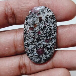 Może przedstawiać: Owalny kamień szlachetny z nakrapianym wzorem czerni, bieli i czerwieni. Kamień ma wypolerowaną powierzchnię i jest trzymany w dłoni. Kamień szlachetny to kamień naturalny.